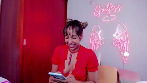 Media: Video of a happy woman with dark hair in buns, wearing a red top, holding a phone, in a room with red door, neon \"Goddess\" sign, and angel wings.