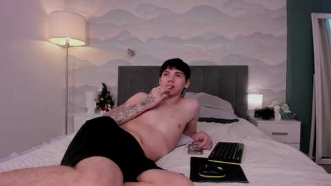 Media: Video of a shirtless young man with tattoos, lying on a bed in black shorts, holding a phone, in a modern bedroom with a white and teal decor.