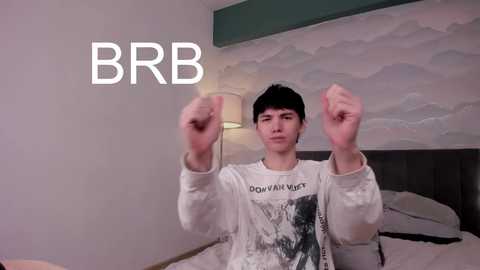 Media: Video of a young man in a white graphic tee, punching the air, in a modern bedroom with a minimalist, cloud-patterned wall and a bedside lamp. \"BRB\" text overlays the image.