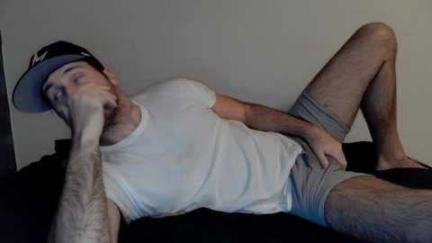 Media: Video of a young, fair-skinned man with a beard, wearing a black cap, white t-shirt, grey shorts, and black socks, lying on a dark bed against a plain wall, hand over his mouth, legs spread, hairy legs.