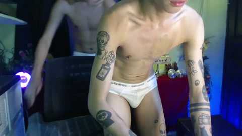 Media: Video of a shirtless, slender, pale-skinned man with tattoos, wearing white Calvin Klein briefs, bending over, in a dimly lit room with a blue light, another person partially visible in the background.