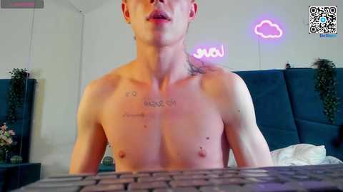 Media: Video of a shirtless, fit, fair-skinned man with tattoos, sitting at a desk in a modern room with a pink neon sign and a blue couch.