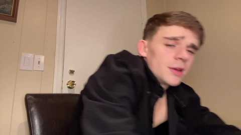 Media: Video of a young man with short brown hair, wearing a black jacket, sitting on a dark leather chair, looking pensive in a beige-walled room with a white door and a light switch.