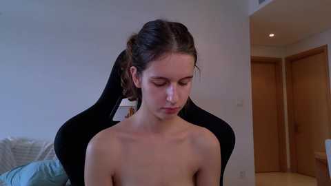 Media: A video of a young, light-skinned woman with dark hair tied back, sitting topless in a black gaming chair, in a sparsely decorated, dimly lit room with wooden doors and a bed in the background.