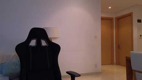 Media: Video of a minimalist bedroom with a black gaming chair facing a white wall, wooden door, and beige carpet.