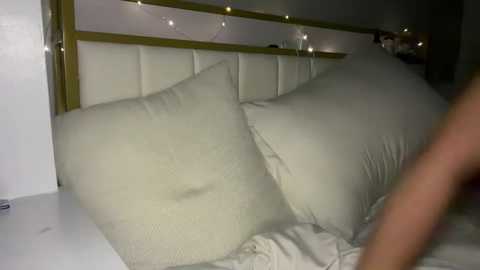 Media: Video of a dimly lit bedroom featuring a white bed with a gold headboard, adorned with a string of fairy lights, and two large, fluffy white pillows.