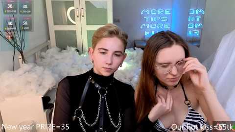 Media: Video of two women in a room with white faux fur and neon signs. One wears black leather harness, the other adjusts glasses.