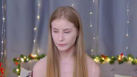 Media: Video of a fair-skinned, blonde-haired woman with a serene expression, wearing a strapless top, in a festive room with string lights and holiday decorations.