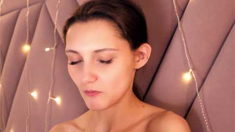 Video of a young woman with light skin and dark hair, wearing minimal makeup, sitting against a soft, mauve, quilted backdrop. Warm, fairy lights create a cozy, intimate atmosphere.