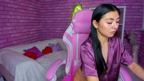 Media: Video of an Asian woman with long black hair, wearing a purple satin robe, sitting in a pink gaming chair in a pink-brick-walled room with a bed, pillows, and decor.