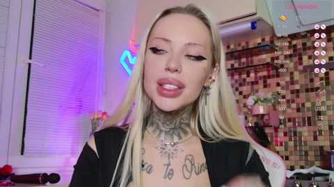 Media: Video of a blonde woman with heavy makeup, tattoos, and piercings, wearing a black top, standing in a colorful, tiled room with neon lights and a TV screen.