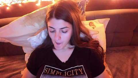 Media: Video of a young woman with long brown hair, wearing a black \"Simplicity\" t-shirt, lying on a beige couch with white pillows, under string lights, eyes closed.