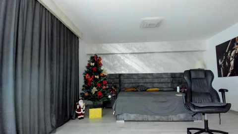 Video of a modern, minimalist bedroom with a grey bed, black curtains, a large Christmas tree adorned with red, white, and green ornaments, and a black ergonomic office chair.