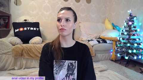 Media: Video of a young woman with long brown hair in a black graphic t-shirt, sitting in a cozy living room with a decorated Christmas tree and plush toys.