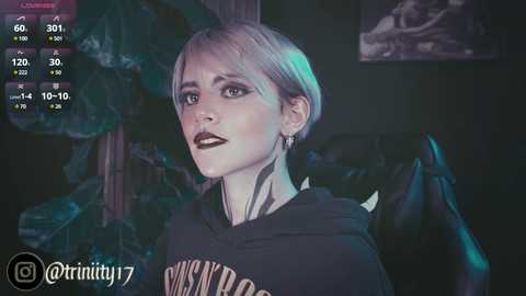 Media: Video of a young woman with short, light purple hair, wearing dark makeup, a black hoodie, and a tattoo on her neck. She is sitting in a dimly lit room with plants in the background.