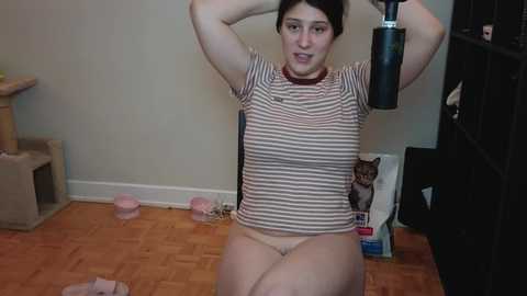 Media: A video of a young woman with fair skin and dark hair, wearing a striped shirt and beige panties, performing an exercise with a kettlebell in a simple, cluttered room with wooden floors and a cat sitting nearby.