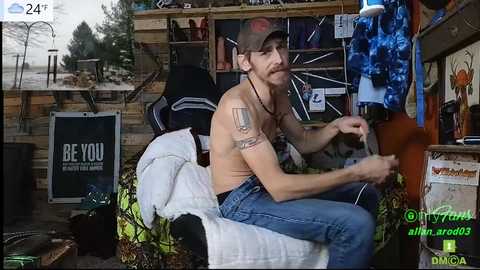 Media: A video captures a shirtless, tattooed man with a beard and cap, sitting in a cluttered room with various objects and a \"BE YOU\" sign.