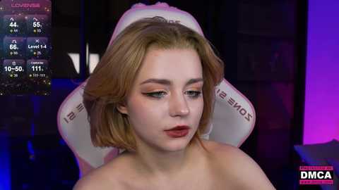 A video of a young woman with short, blonde hair, wearing makeup, sitting in a gaming chair with a purple background. She appears to be in a livestream.