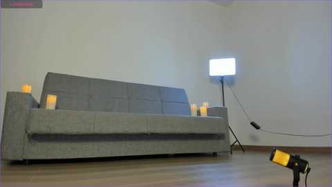 Media: Video of a minimalist living room with a grey, L-shaped sofa, lit by a bright, white rectangular lightbox; warm-toned candles scattered on the sofa, wooden floor, and a yellow flash on the floor.