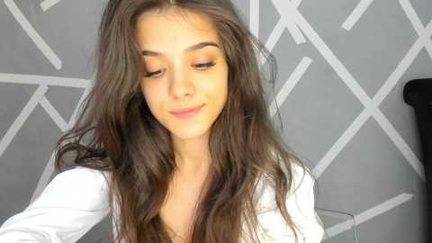 Media: Video of a young woman with long, wavy brown hair, fair skin, and closed eyes. She wears a white top. Background features geometric, grey-patterned wallpaper.