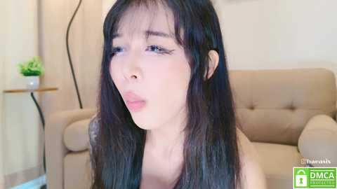 Media: Video of an Asian woman with long black hair, pale skin, and a light beige couch in the background. She has a neutral expression, slightly sticking out her tongue.