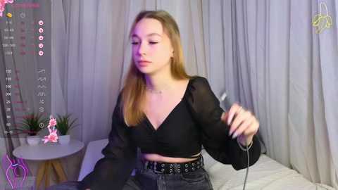 Media: Video of a young Caucasian woman with fair skin and long blonde hair, wearing a black crop top and high-waisted jeans, sitting in a dimly lit room with a pink flower on a table and gray curtains.