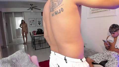 Media: Video of a shirtless man with a tattoo on his back, wearing white pants, standing in a modern, minimalist living room. A woman sits on the floor reading a book.