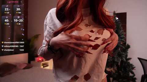 Media: A video of a red-haired woman in a sheer, white, geometric-patterned top, covering her breasts, in a dimly lit room with a Christmas tree in the background.