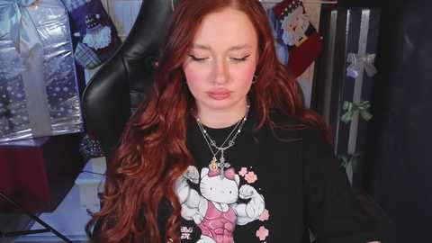 Media: Video of a young woman with long, wavy red hair, wearing a black Hello Kitty shirt, sitting in a black gaming chair, surrounded by festive wrapping paper.