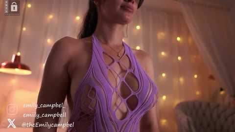 Video of a woman in a revealing, purple crochet halter top, standing in a softly lit, cozy room with string lights and white drapes.