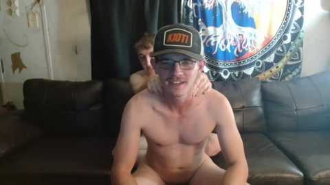 Media: Video of two shirtless men on a dark brown leather couch; one wears glasses and a baseball cap, the other has a beard, both smiling. Background includes a colorful tapestry and a door.