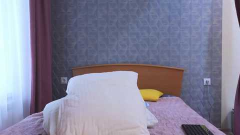 Media: Video of a neatly made bed with a white pillow, a yellow pillow, and a pink blanket, against a textured blue-gray wallpaper background.