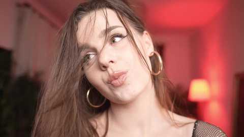 Media: Video of a young woman with long brown hair, wearing large gold hoop earrings, making a pouty face, in a softly lit, red-hued room.