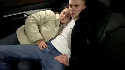 Media: Video of a young couple cuddling in a car, the woman with light hair in a beige puffer jacket, the man in a white shirt, both looking relaxed and close.