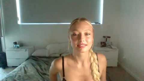 Media: Video of a blonde woman with braided hair, wearing a black tank top, standing in a modern, minimalist bedroom with white walls, a bed, and gray bedding.