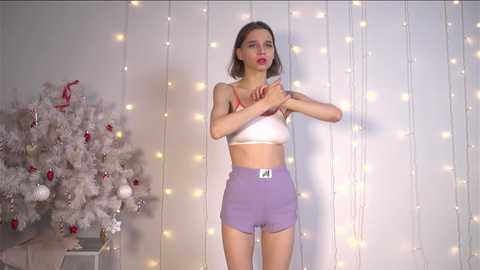 Media: Video of a slender, fair-skinned young woman with shoulder-length brown hair, wearing a white sports bra and purple shorts, dancing in front of a string-lit white wall and a decorated Christmas tree.