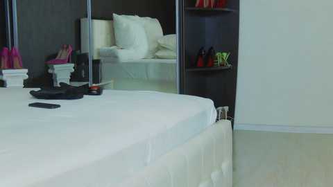 Media: A video of a minimalist, modern bedroom with a white bed, black wall, and mirrored wardrobe. The room features a tufted headboard, a black smartphone, and red shoes on a shelf.