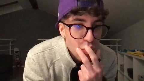 Media: Video of a young man with curly hair, wearing a purple baseball cap, glasses, and a grey hoodie, sitting and touching his nose, indoors with dim lighting.