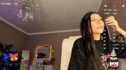 Media: Video of a woman with long black hair, wearing a black sweater, looking at a smartphone displaying a video game. Background includes a Christmas tree, a mirror, and a white couch in a cozy room.