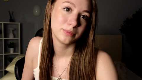 Media: Video of a young woman with long, straight, light brown hair, fair skin, wearing a white tank top, sitting indoors. She has a neutral expression, with a minimalistic background featuring a white shelf with decorative items.