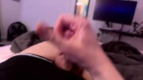 Media: A blurred video of a person lying on a bed, with a clenched fist raised, wearing black underwear. Background includes a TV, a fan, and a dark room with white walls.