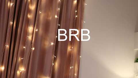 Media: Video of a room with a maroon curtain adorned with warm, glowing fairy lights. The text \"BB\" is centered in white letters, suggesting a brand or product. The background is a soft, neutral beige wall.