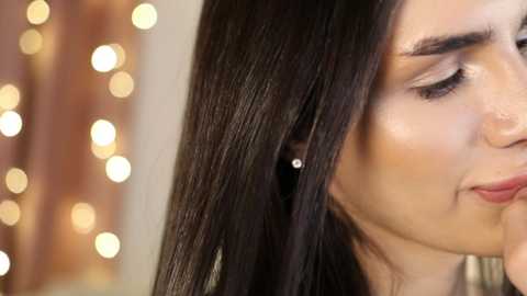 Media: A close-up video of a young woman with long, straight, dark brown hair, fair skin, and subtle makeup. She wears a single small, sparkly earring and is seen from the side, gazing downward. The background features a blurred, warm-toned, bokeh effect.