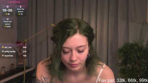 Media: Video of a young woman with fair skin and shoulder-length, wavy green hair, wearing a white, spaghetti-strap top. Background shows a potted plant and dimly lit room with grey curtains.