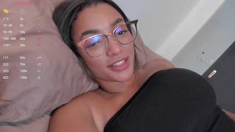 Media: Video of a light-skinned woman with glasses, black hair, and a black top, lying on a bed, smiling. Calendar and digital clock visible in the background.