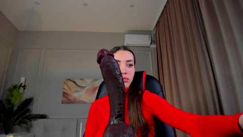 Media: A video of a young woman with long, dark hair, wearing a bright red jacket, sitting in a modern living room with beige walls and a floral painting. She has a large, dark brown boot covering her face, creating a humorous and surreal scene.