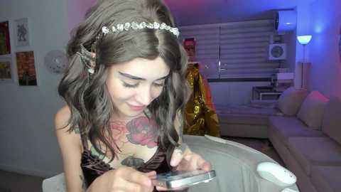 Media: A video of a young woman with long brown hair and a floral tattoo on her chest, wearing a black dress, playing with a smartphone in a modern living room with a white couch, a TV, and a ceiling fan.