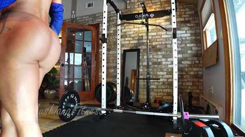 Media: Video of a muscular woman, topless with a blue sports bra, lifting weights in a rustic gym with exposed brick walls and large windows.