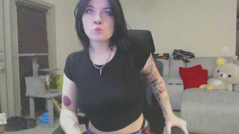 Media: Video of a tattooed woman with black hair, wearing a black crop top, making a kissy face in a cluttered living room.