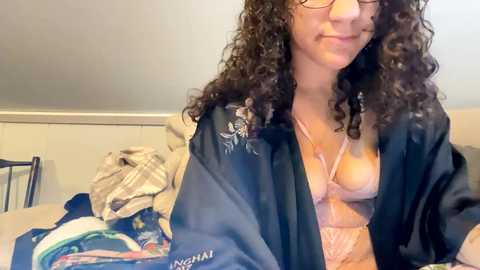 Media: Video of a light-skinned woman with curly hair, wearing glasses, a black robe, and a lacy pink bra, sitting on a bed with messy clothes in a dimly lit room.